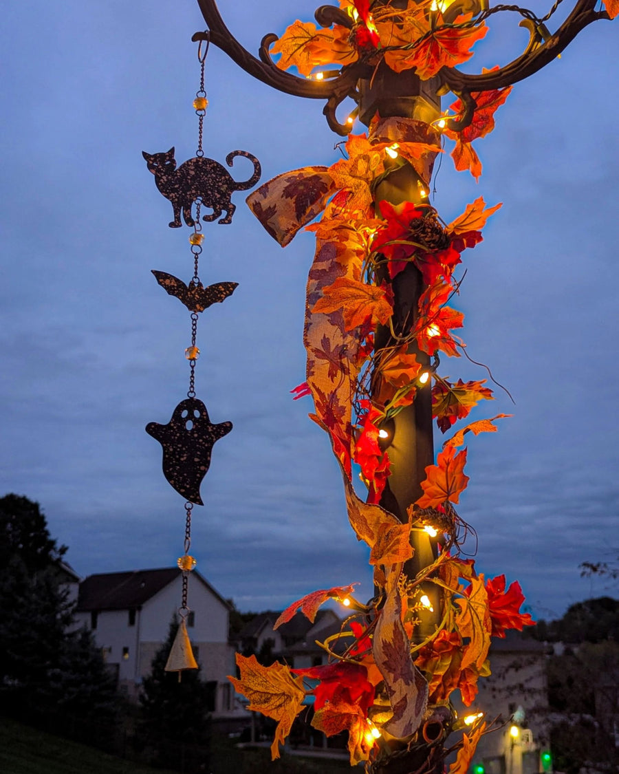 Halloween wind chime with cat, bat, and ghost cutouts hanging beside an autumn leaf garland with lights outdoors.