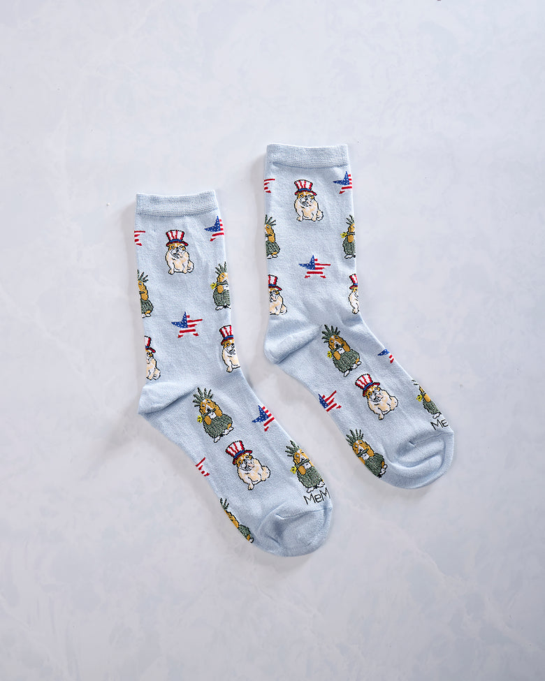 Two light blue crew socks displayed side by side with repeated patriotic dogs, pineapple-like figures, and American flag motifs.