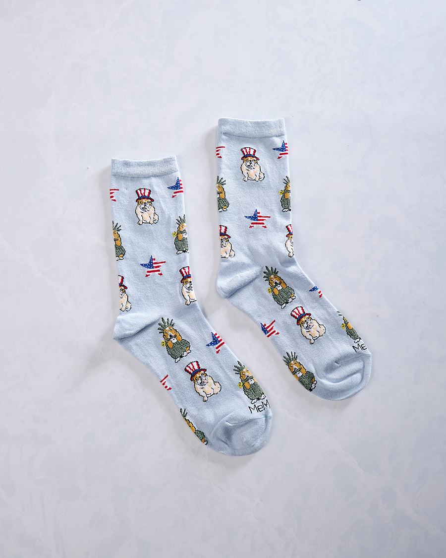 Two light blue crew socks displayed side by side with repeated patriotic dogs, pineapple-like figures, and American flag motifs.