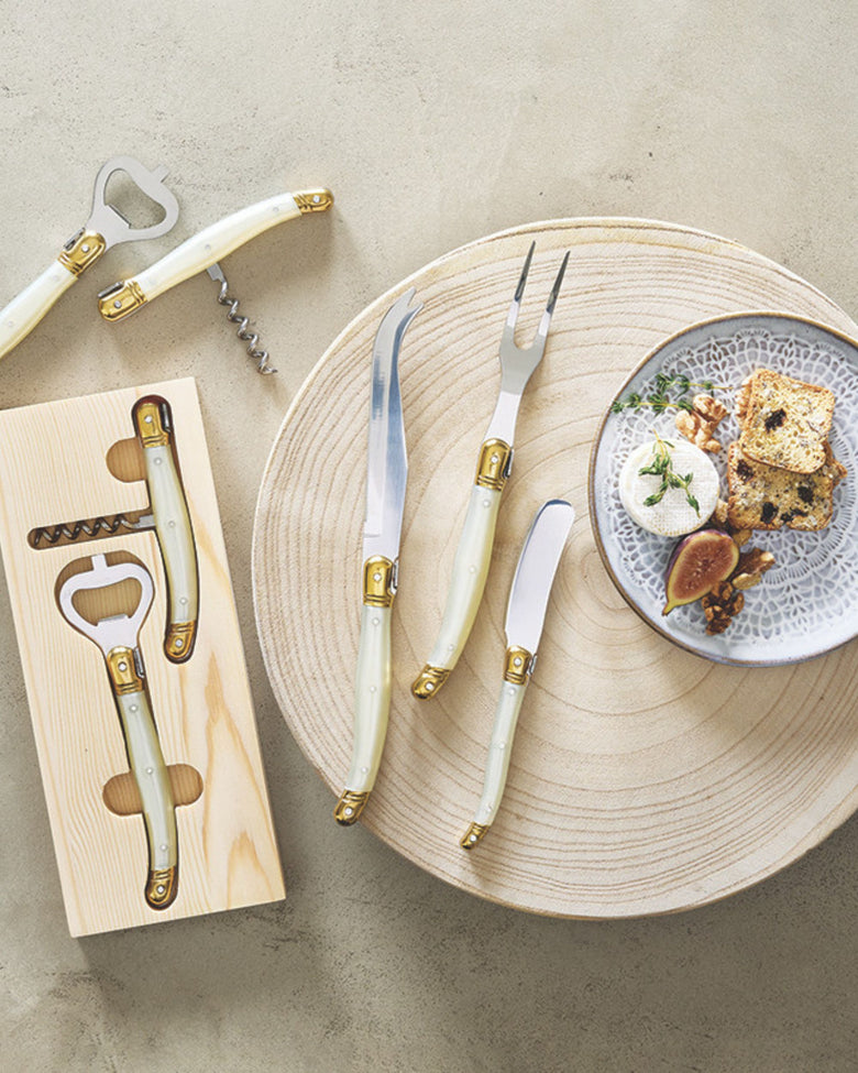 Cheese knife set arranged on a round wooden board beside a plate of cheese and bread, with the tray and bottle opener visible.