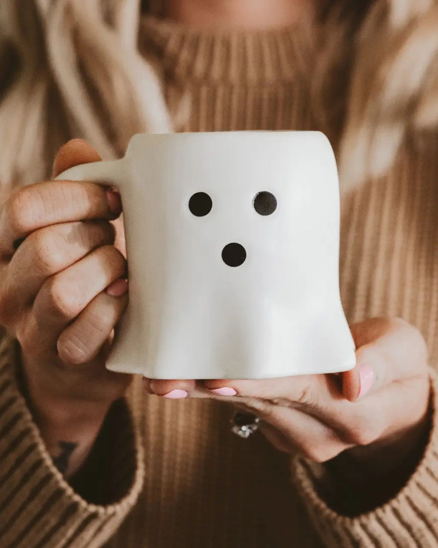 White ghost-shaped coffee mug with a three-dot face, held in a person's hands.