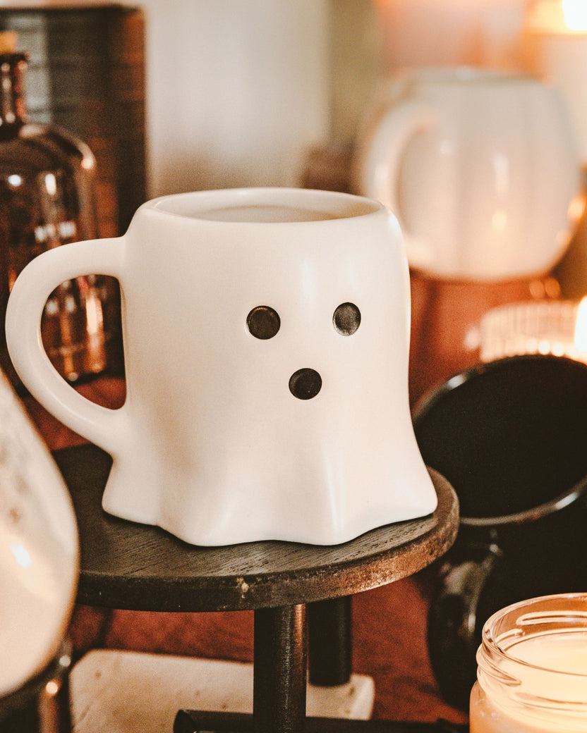 White ghost-shaped mug displayed on a small round wooden stand among seasonal decor and candles.